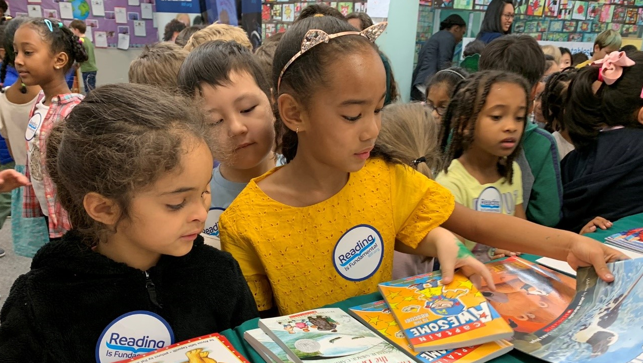 Children pick out books at an event organized by Reading Is Fundamental and Nestle. A girl in a yellow shirt with a golden headband stands at the front of the table, reaching out to pick a specific title. Two nearby children look over her shoulder at the spread of books.