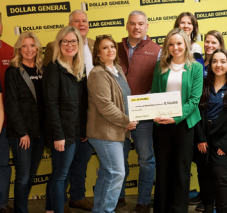 Fourteen people stand together, including members of the team from Reading is Fundamental and Dollar General. Two people hold an oversized check in the center of the group.