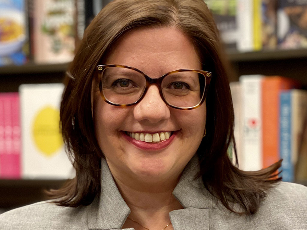 Jackie DeLeo, the Chief Merchandising Officer of Barnes & Noble, smiles at the camera. She wears tortoiseshell glasses and a gray jacket. She is standing in front of a shelf of books.